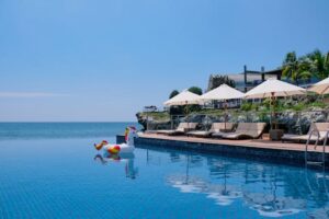 A tranquil beachside infinity pool with sunbeds, umbrellas, and a playful unicorn float under the summer sun.